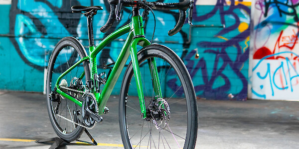 A custom-designed carbon Plane Frameworks bicycle in bright green, viewed on a three-quarter angle, a graffiti-covered wall in the background