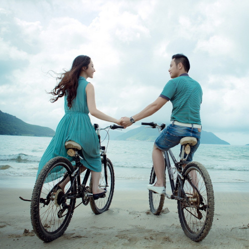 A couple sitting on bicycles at the beach, holding hands and looking at each other