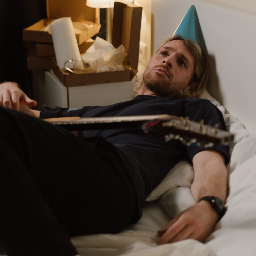A young bearded man wearing a party hat, lying across a bed with a guitar in his lap, looking despondent