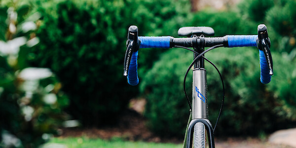 A custom-built Bossi Summit titanium road bike, viewed from the front in a garden setting, the hand-painted head tube decal and matching blue handlebar tape visible