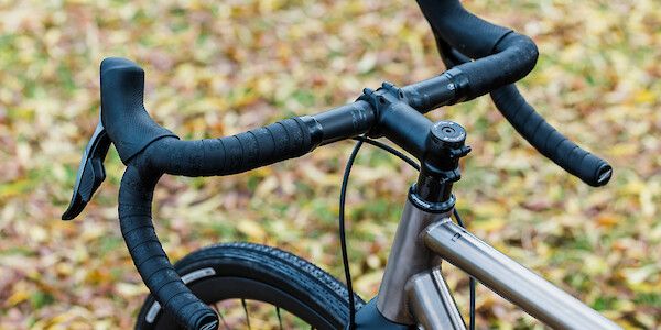 Handlebar detail on a titanium Bossi Grit gravel bike, viewed from the left at an angle, against a background of leaves
