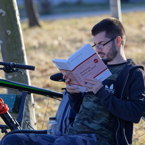 A man with glasses sitting on a bench and reading a book, next to a bicycle