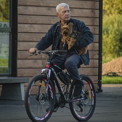An elderly gentleman riding a bicycle, holding a terrier in one arm. Both dog and rider look displeased.