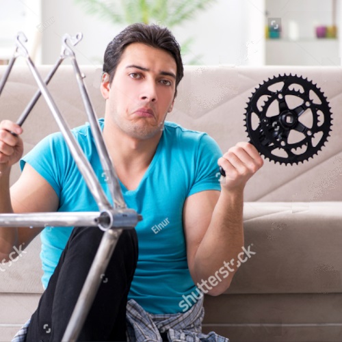 A man sitting in front of a couch with a disassembled bicycle, shrugging his shoulders