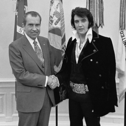 Black and white photo of former president Richard Nixon in the White House, shaking hands with Elvis Presley. Both are facing the camera. Elvis looks slightly dazed.