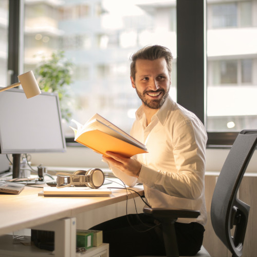 A bearded man sitting in front of a computer and holding a book. He is smiling at someone over his shoulder.