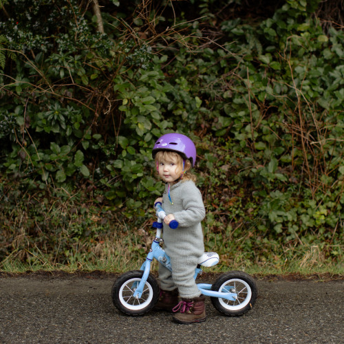 A small girl on a balance bike in front of a hedge. She is giving the camera side-eye.