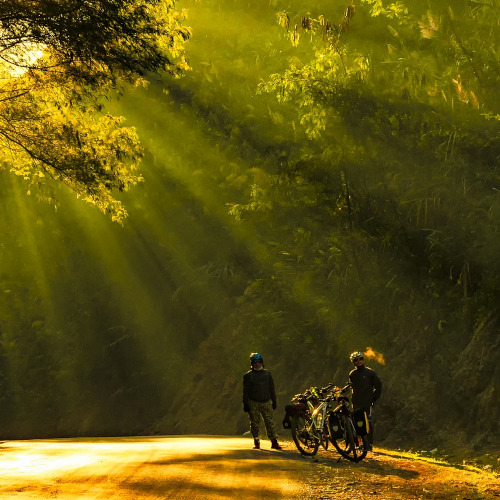 Two cyclists with loaded bikes, standing roadside in a streaming pool of golden sunlight. It looks cold out there, despite the sun.
