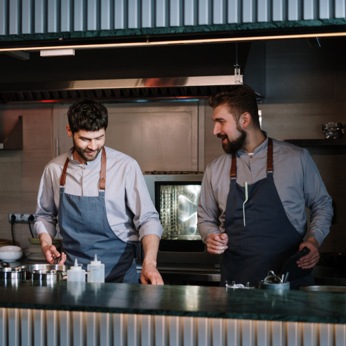 Two chefs working at an open window and having a discussion. They are both bearded, with leather-strapped aprons. We don't know what their food is like.