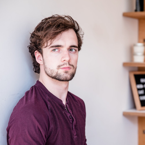 Head-and-shoulders shot of a young man with a curated beard, leaning against a wall inside a shop and despondently staring into the middle distance.