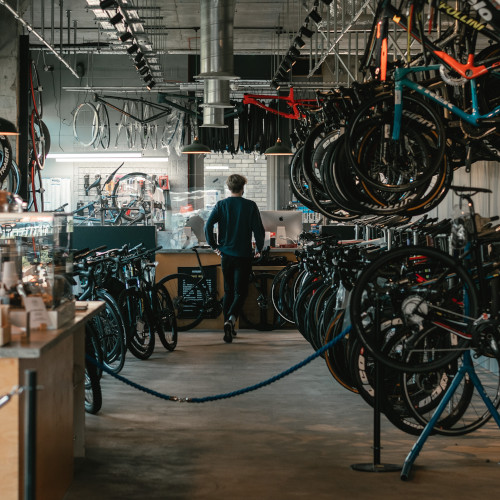 A young man walking away into a neat and tidy bicycle workshop, rows of bikes on the floor and also hanging up.