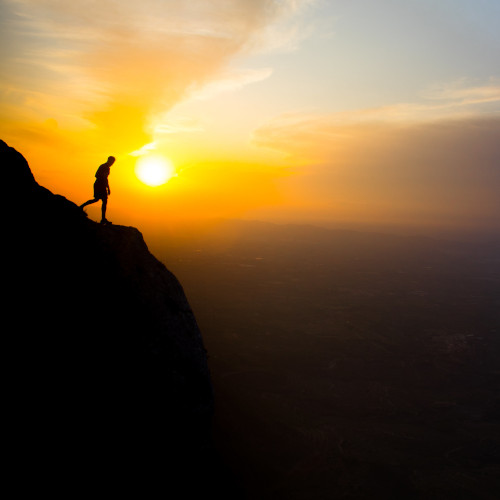 A man silhouetted on the edge of a cliff against a sunset, looking down over the edge