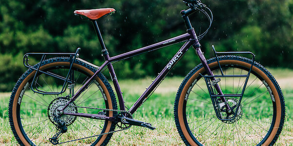 A custom-built Surly Karate Monkey bicycle in Eggplant, complete with Surly racks and Brooks saddle, standing in a field of green.