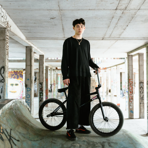 A young man dressed in baggy black, standing with a BMX inside a skate park.