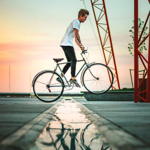 A young man jumping a street bicycle over a puddle, his reflection in the water in the foreground.