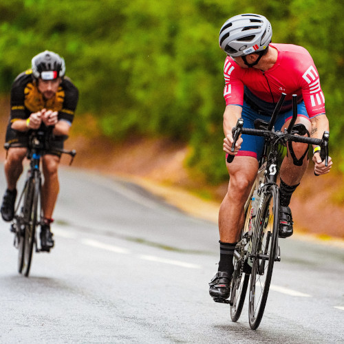 Two road cyclists coming down a hill during a race; the one in front is looking back to the second one.