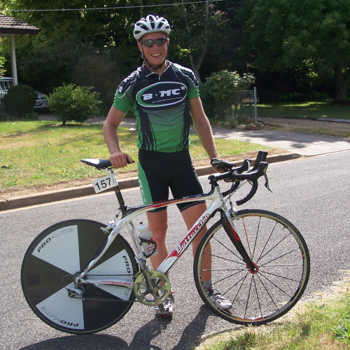 A young male cyclist wearing BMCR lycra kit, posing with a Bottecchia road bike with a time trial disc wheel in the back.
