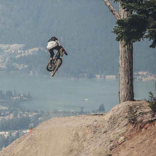 A mountain biker mid-air over a large jump, possibly dismounting. He's facing away from the camera; there are hills and a city in the distance.