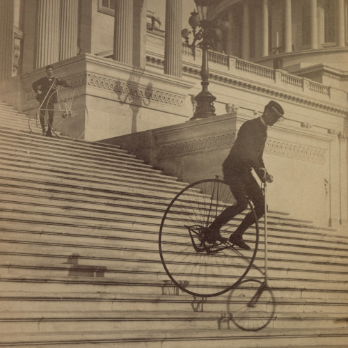 An old photograph of a man cycling a penny farthing bicycle down the steps of a Greek-style building.