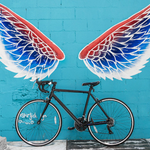 A black bicycle leaning against a blue wall, a mural of wings painted above.