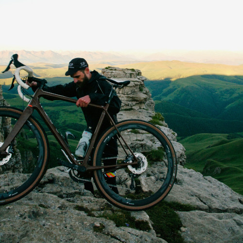 A man manouvering a bicycle over some rocks, in front of a mountain range