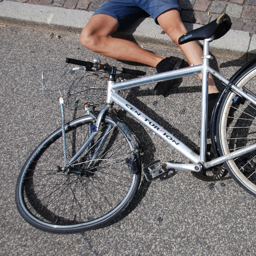 A silver bicycle with a crushed front wheel lies on the ground. The legs of its rider can be seen, splayed on road behind the bike.