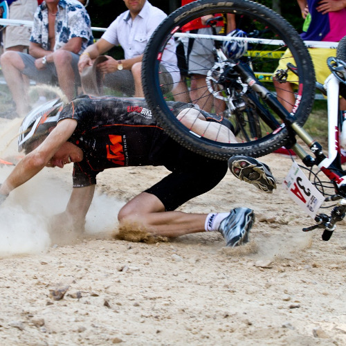 A mountain biker crashing his bike mid-race. He is tumbling onto the ground, tangled with his bike, watched by spectators.