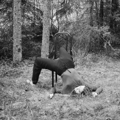 A black and white photo of a woman in a forest, half-lying on the ground with her legs tangled in a chair.