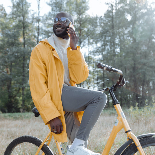 A man sits on a bike, trees in the background, talking on a phone. His bike, jacket and phone are a matching yellow.