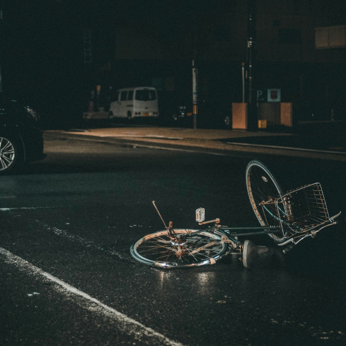 A crashed bicycle lies in the middle of the street, a car visible in the background. It is a dark and stormy night.