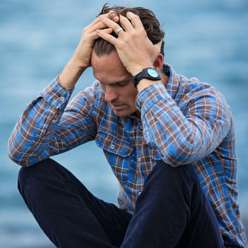 A young man sits by a body of water, his head in his hands in despondence.