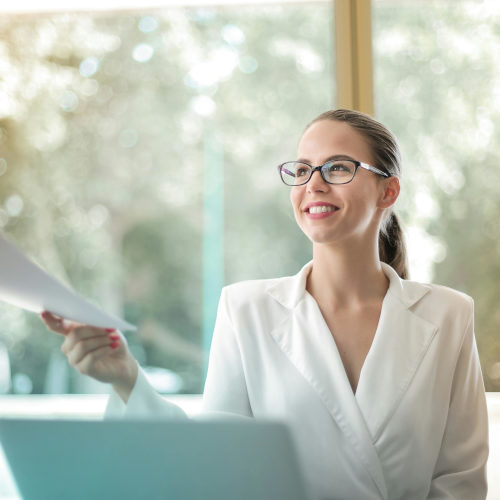 A young woman at a desk hands a piece of paper to an unseen person.