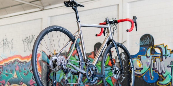 A Bossi Grit titanium gravel bike in a custom build, viewed from ground level, a graffitied wall visible in the background