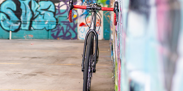 Front view of a custom-built titanium Bossi Grit gravel bike with bright red handlebar tape, leaning against the wall of a graffitied car park