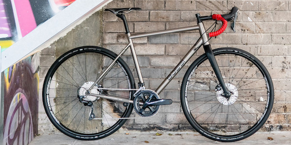 A custom-built titanium Bossi Grit gravel bike with bright red handlebar tape, leaning against a brick wall, graffiti visible in the foreground