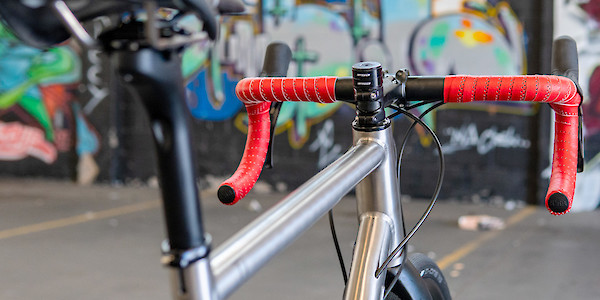 A Bossi Grit titanium gravel bike with bright red handlebar tape, viewed from the back near the seat post, a graffitied wall visible in the background