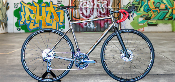 A custom-built Bossi Grit titanium gravel bike, viewed from the side, a colourful graffitied wall in the background