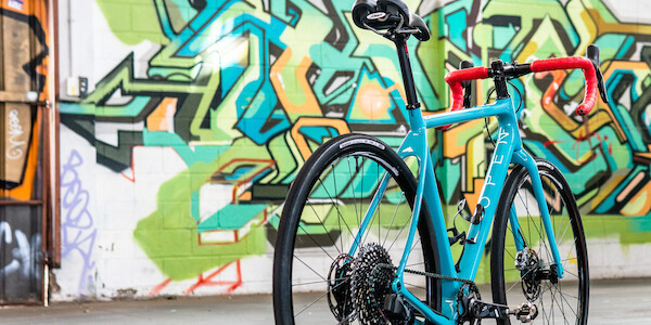 A blue Open Cycles WiDe custom-built gravel bike, viewed at an angle from the rear, in front of a colourful graffitied wall