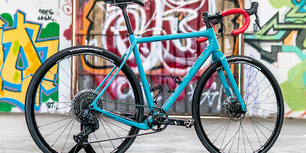 A custom-built blue Open Cycles Wi.De gravel bike with red bar tape, a colourful graffitied wall in the background