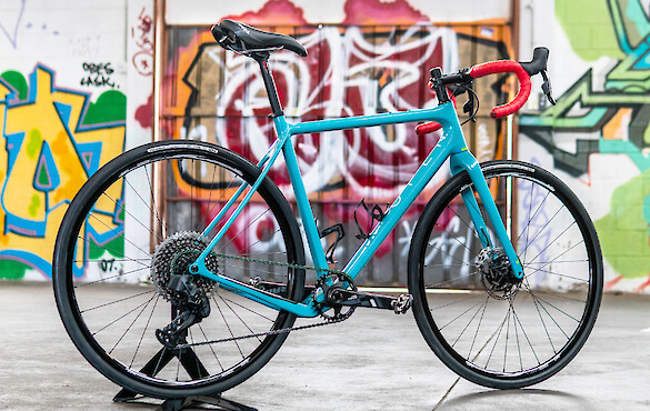 A custom-built blue Open Cycles Wi.De gravel bike with red bar tape, a colourful graffitied wall in the background