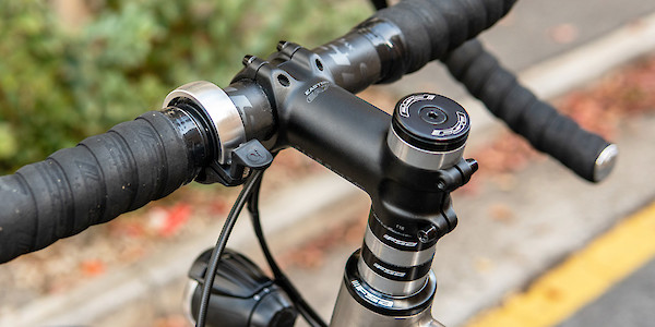 Headset spacer and handlebar detail on a Bossi Grit titanium bike, viewed from the back and to the left. A brass Knog Oi bell is also visible on the bars.