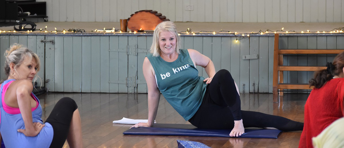 Remedial massage therapist Holly Hicks teaching a stretch class in a community hall, fairy lights on the stage behind her