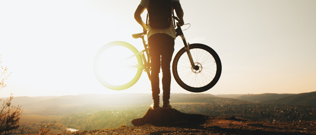 A mountain biker, facing away from the camera, holding up a mountain bike at waist height and looking over the horizon.