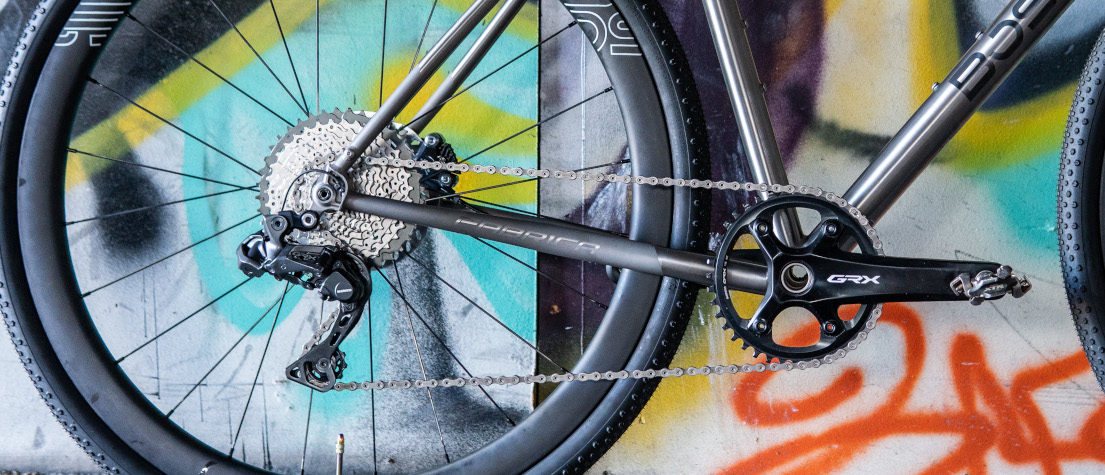 Drivetrain/gear component detail on a titanium Bossi bike which is leaning against a graffitied wall