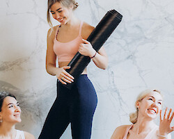 Women in fitness attire, talking inside a studio. One is holding a yoga mat.