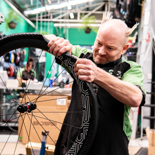 A professional bike mechanic at Bio-Mechanics Cycles & Repairs fitting a tyre to a bicycle wheel. It's a tight fit. His forearm muscles are impressive.
