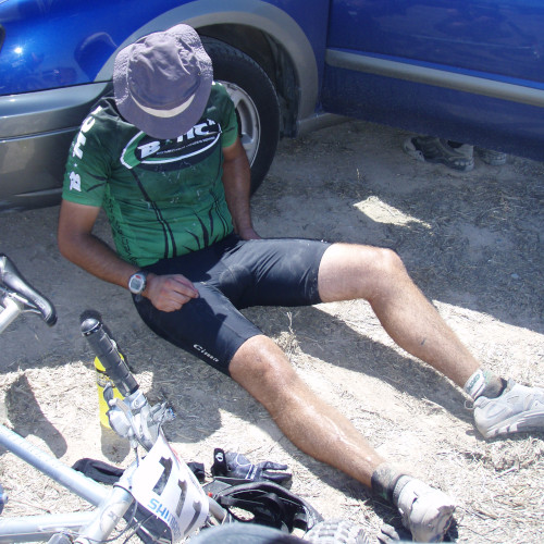 A cyclist in a BMCR jersey, sitting on the ground and leaning against a car, his hat over his face.