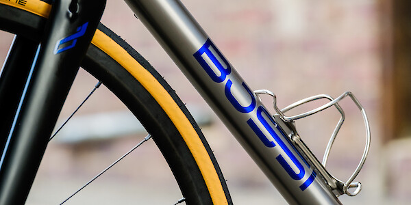 Close-up of blue custom-painted frame and fork decals on a titanium Bossi Summit bicycle, contrasting with the tan sidewall of the front tyre