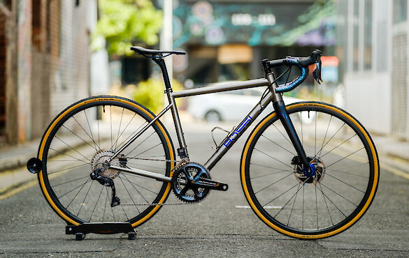 A custom-built titanium Bossi Summit drop-bar bicycle with blue highlights, shot in a city alleyway