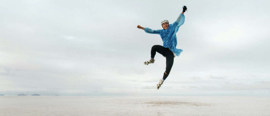 A young man wearing cycling gear and a rain jacket, jumping for joy on a deserted beach.